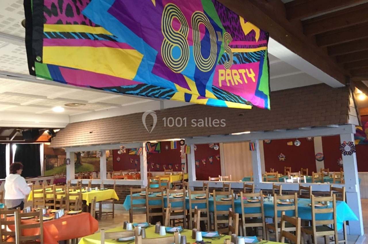 Salle décorée pour une fête sur le thème des années 80, avec tables colorées et une bannière ’80's Party’.
