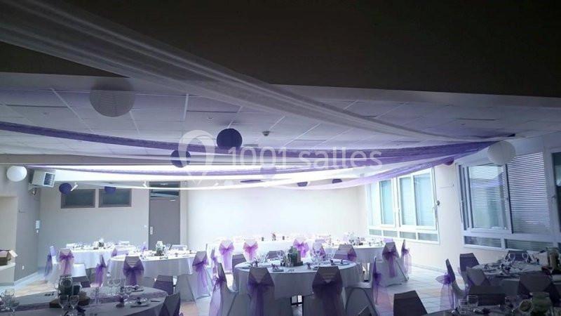 Salle de réception décorée avec des nappes blanches, des chaises ornées de nœuds violets et des guirlandes suspendues.