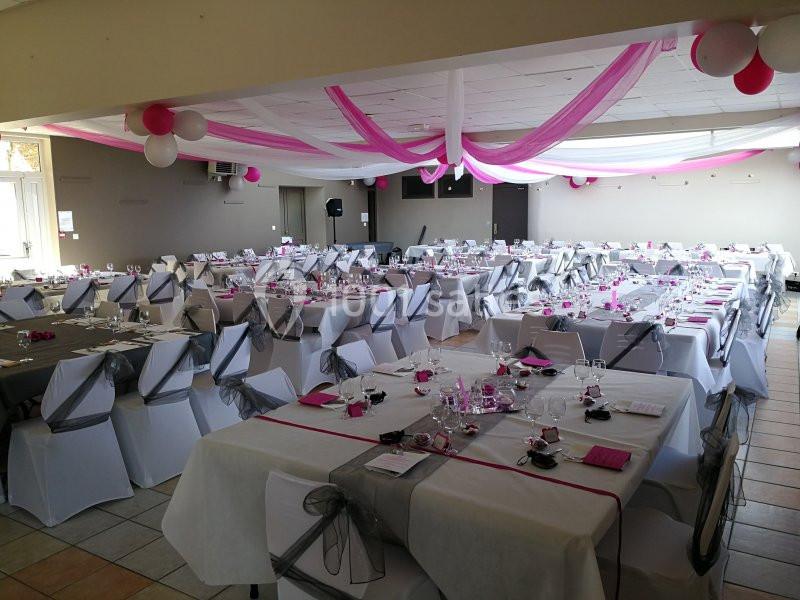Salle de réception décorée avec des nappes blanches, des rubans gris et roses, et des ballons suspendus au plafond.
