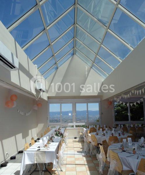 Salle lumineuse avec verrière, tables décorées pour un événement, chaises en bois et vue dégagée sur l'extérieur.