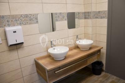 Deux lavabos blancs sur un meuble en bois avec miroirs, distributeur de savon mural et poubelle dans une salle de bain…