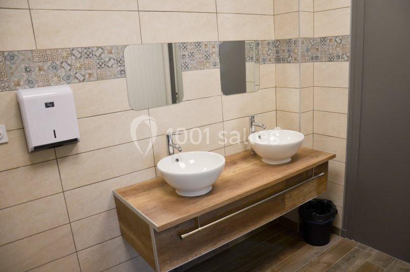 Deux lavabos blancs sur un meuble en bois avec miroirs, distributeur de savon mural et poubelle dans une salle de bain…