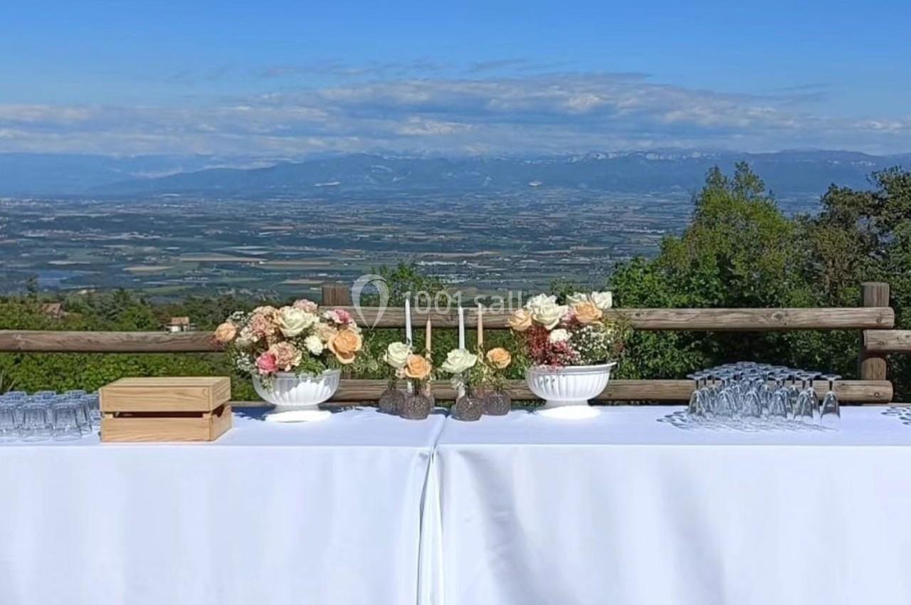 Table dressée en extérieur avec nappes blanches, fleurs, bougies et verres, offrant une vue panoramique sur la vallée.
