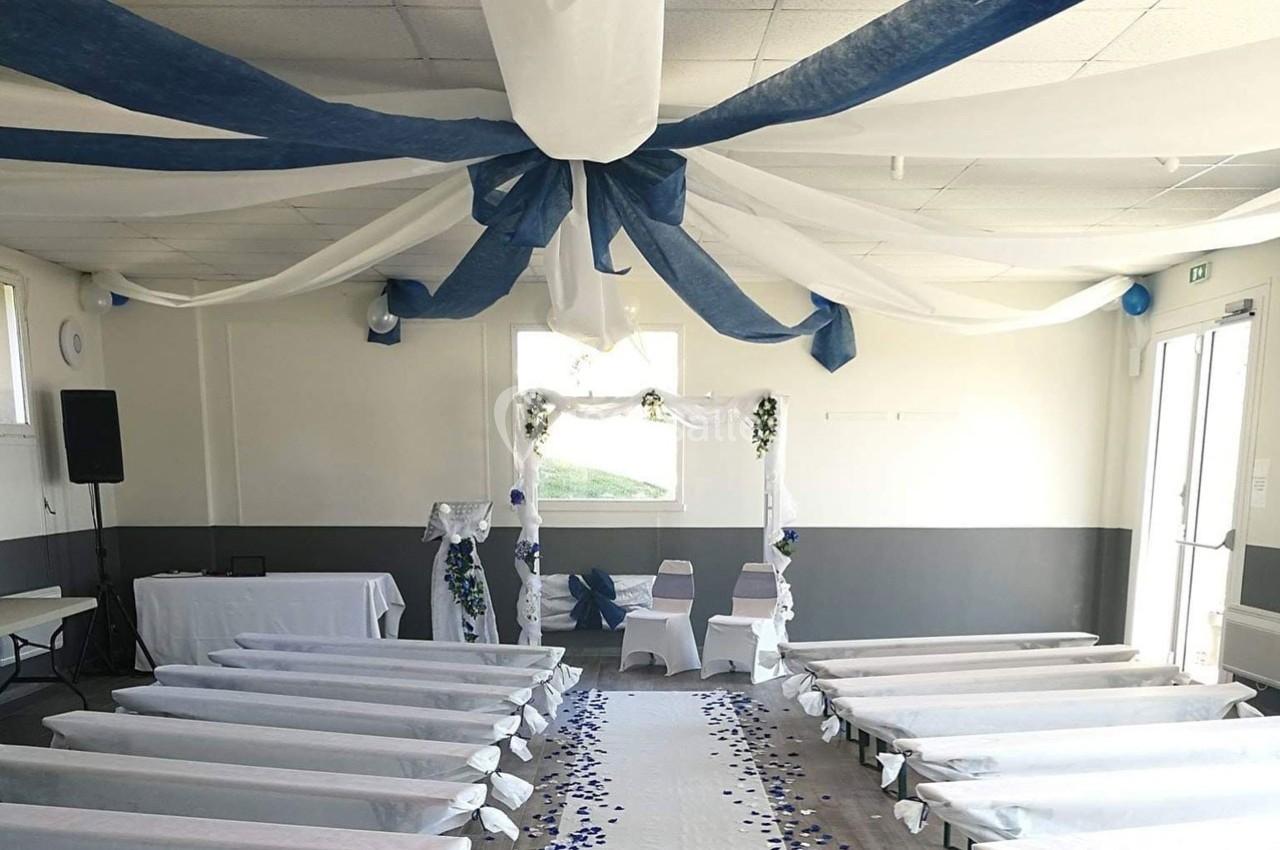Salle décorée pour une cérémonie, avec allée centrale ornée de pétales bleus, chaises blanches et arches fleuries.