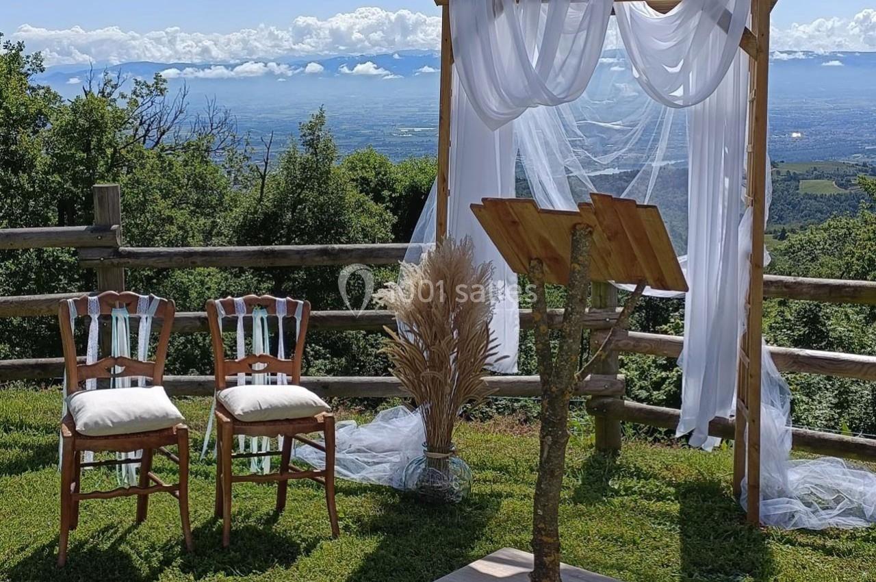 Arche en bois décorée de voilages blancs, deux chaises et pupitre sur une pelouse avec vue sur un paysage vallonné.