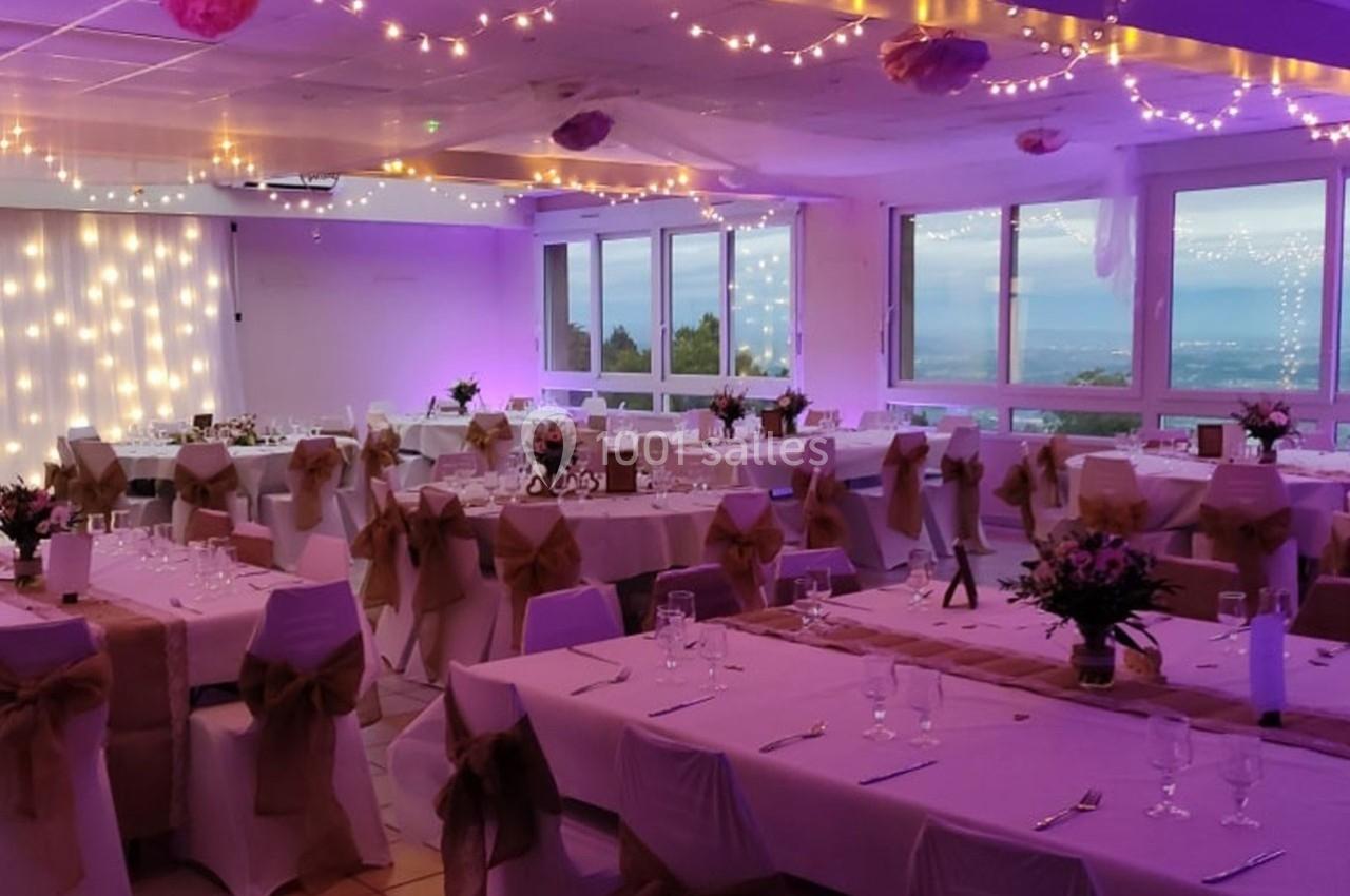Salle de réception décorée avec des guirlandes lumineuses, tables dressées et vue sur un paysage extérieur.