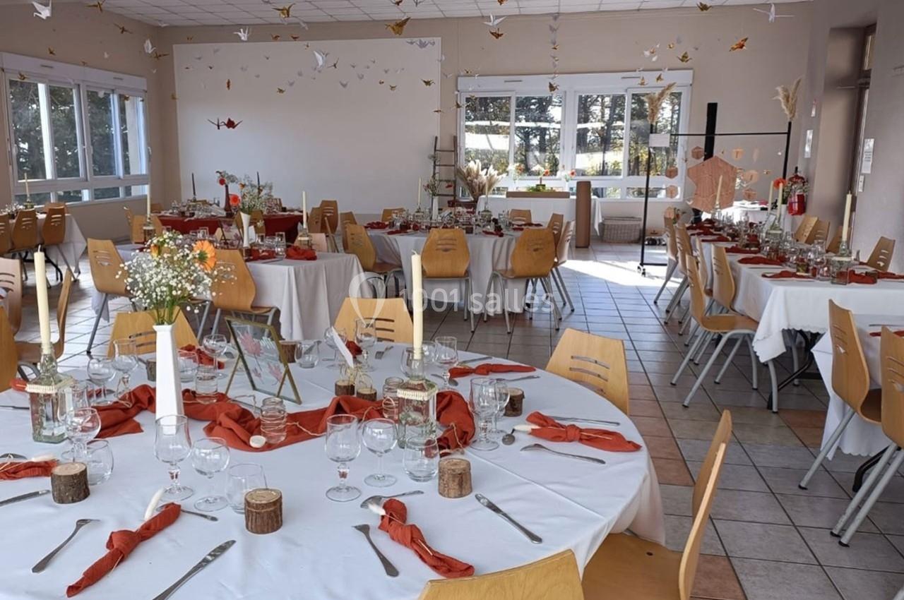 Salle décorée pour un événement avec tables rondes dressées, nappes blanches, serviettes orange et pliages en papier…