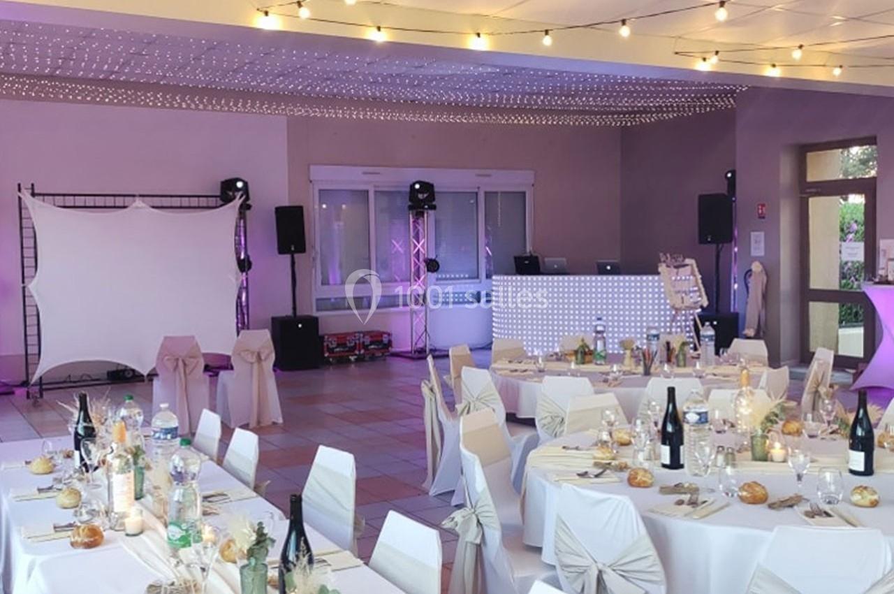 Salle de réception décorée pour un événement, avec tables dressées, chaises ornées et éclairage festif.