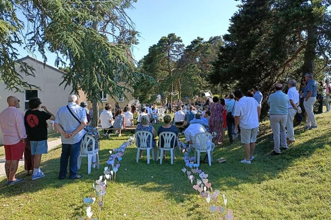 Groupe de personnes assises et debout lors d'une cérémonie en extérieur, entourées d'arbres et de décorations au sol.