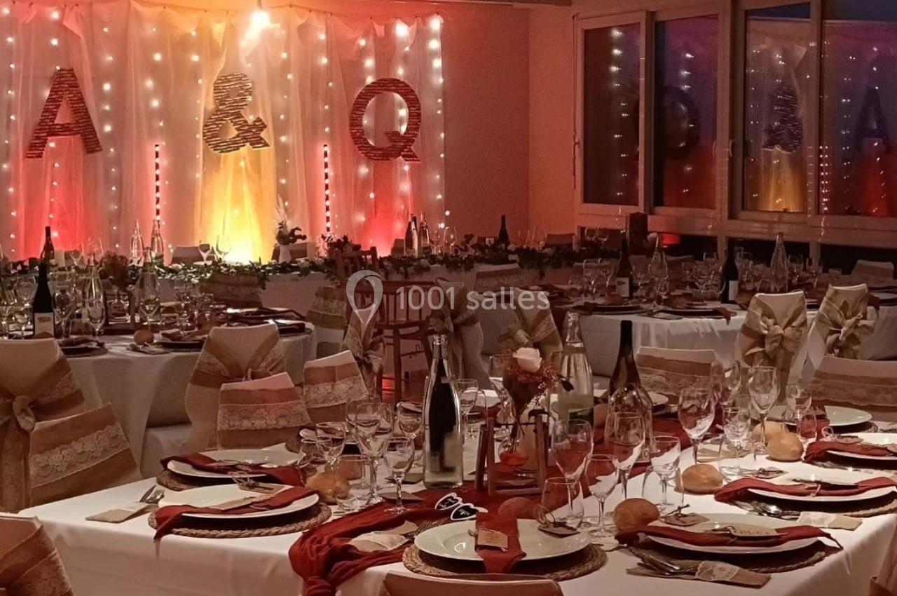 Salle de réception décorée avec des tables dressées, nappes blanches, rubans beiges et lumières chaleureuses en arrière-plan.