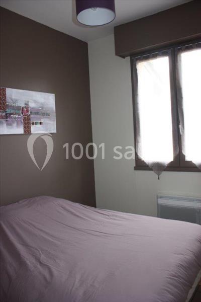 Chambre avec un lit recouvert d'une housse mauve, un mur brun avec un tableau et une fenêtre partiellement voilée.