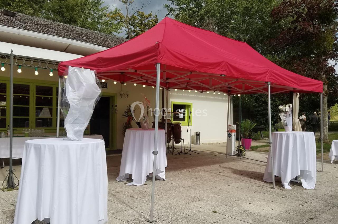 Tente rouge installée en extérieur avec des tables hautes drapées de blanc, devant un bâtiment avec des guirlandes…