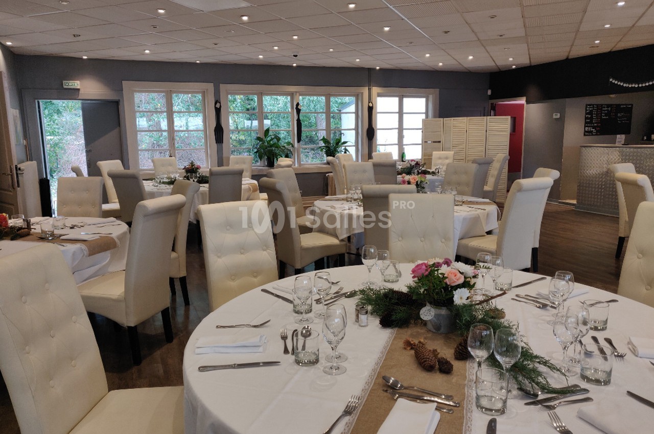 Salle de réception avec tables rondes dressées, nappes blanches, décorations florales et grandes baies vitrées.