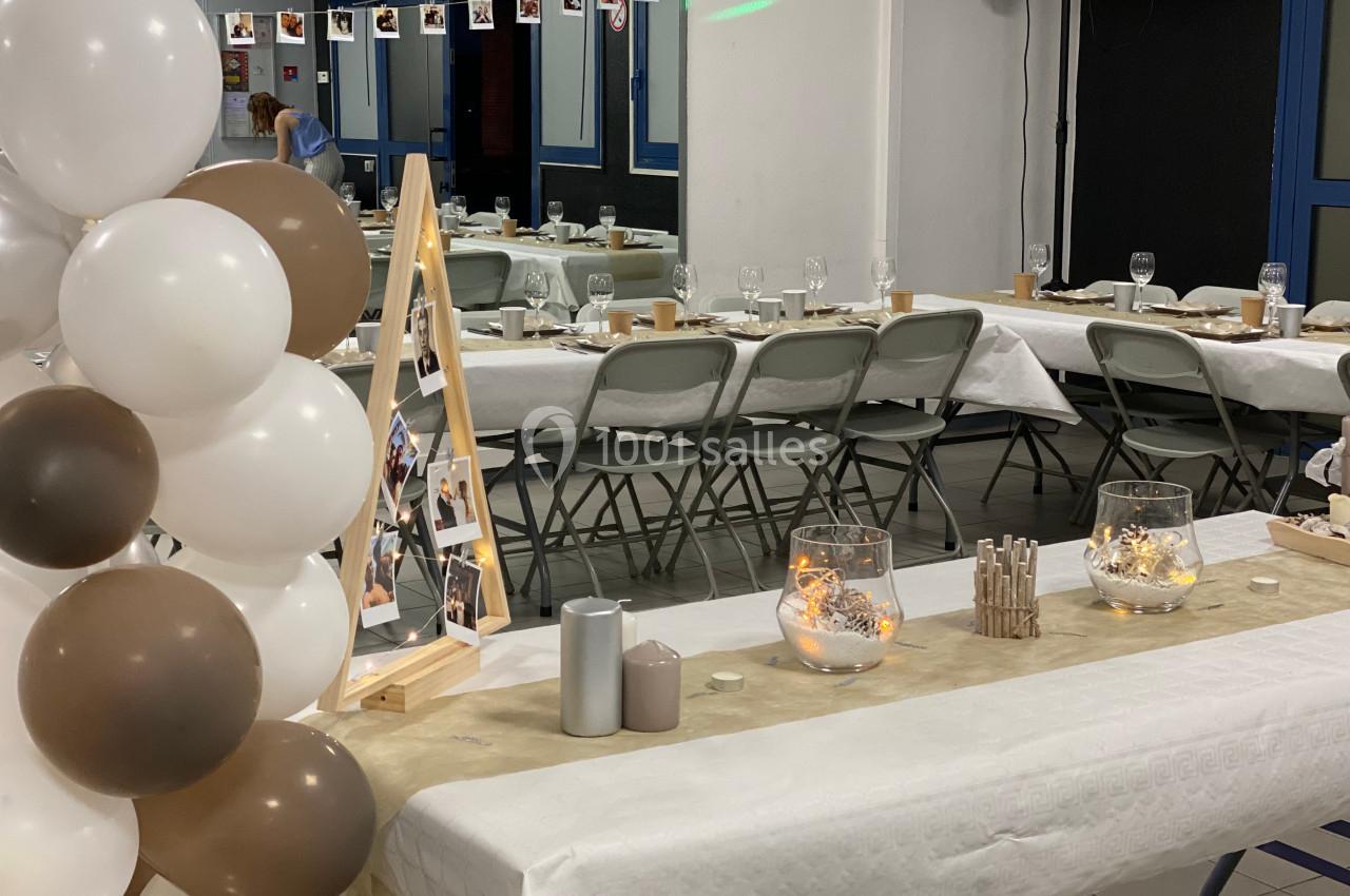 Salle décorée pour un événement avec tables dressées, ballons blancs et marron, et guirlandes lumineuses.