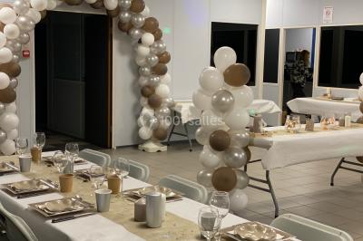 Salle décorée pour un événement avec des arches et colonnes de ballons beige et blanc, tables dressées avec vaisselle.