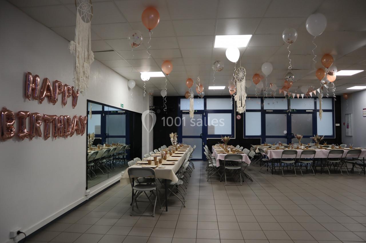 Salle décorée pour un anniversaire avec tables dressées, ballons suspendus et guirlandes dans des tons pastel.