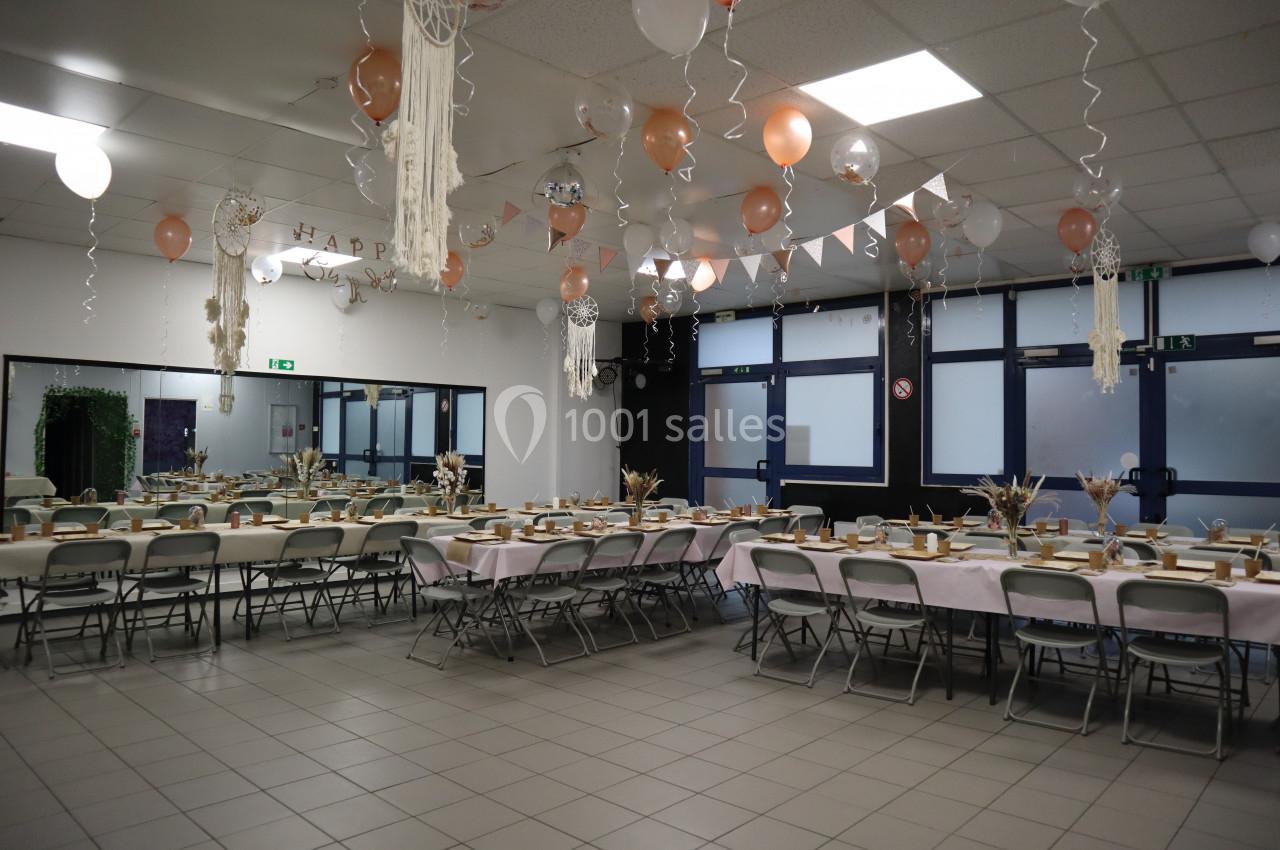 Salle décorée pour un événement avec tables dressées, ballons suspendus et guirlandes dans un espace lumineux.