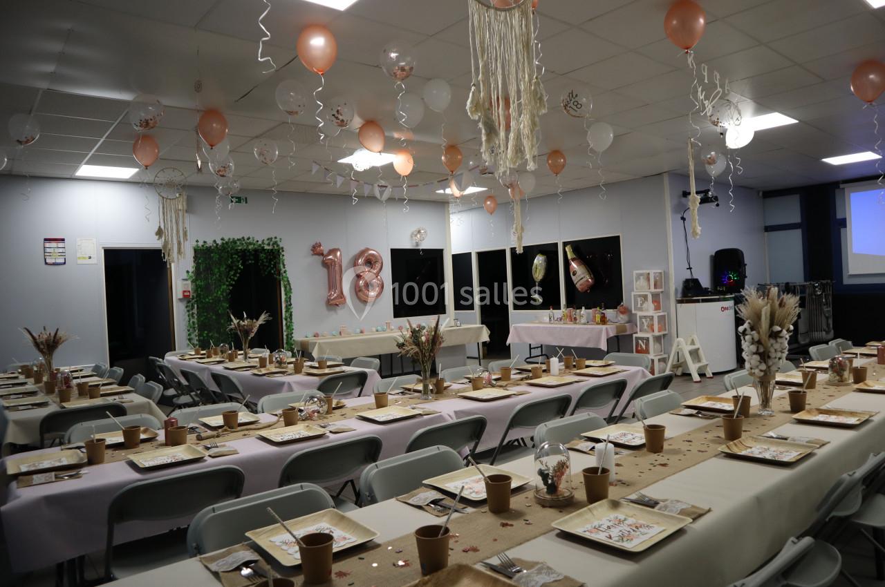 Salle décorée pour une fête avec des ballons, des tables dressées et une arche végétale.