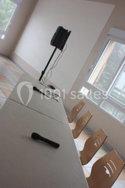 Salle lumineuse avec une table équipée de microphones, des chaises en bois alignées et un haut-parleur sur pied.