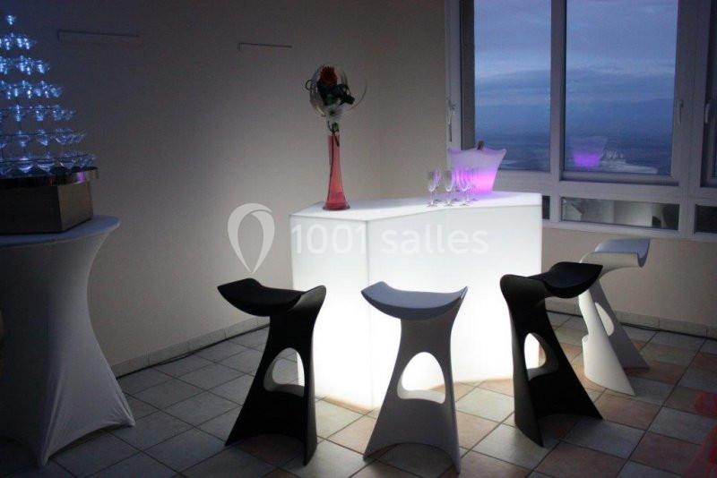 Bar lumineux blanc entouré de tabourets noirs et blancs dans une pièce avec vue sur une fenêtre en soirée.