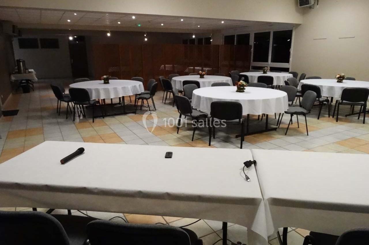 Salle de réception avec tables rondes dressées, chaises disposées autour et une table équipée de micros au premier plan.