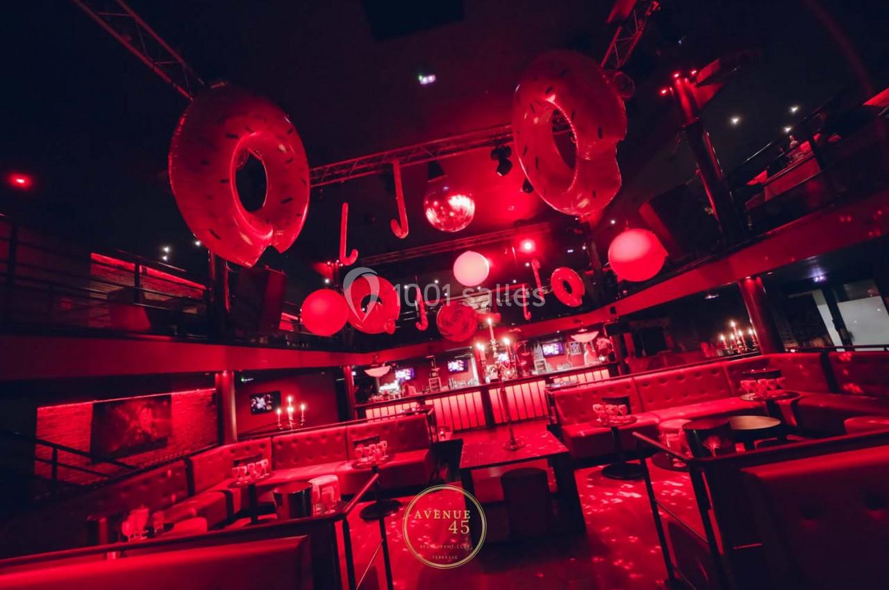 Salle de fête avec éclairage rouge, décorations suspendues et tables entourées de banquettes.