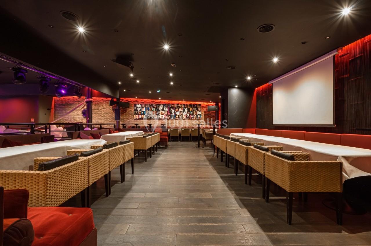 Salle de restaurant avec tables alignées, chaises en osier, éclairage tamisé et bar en arrière-plan.