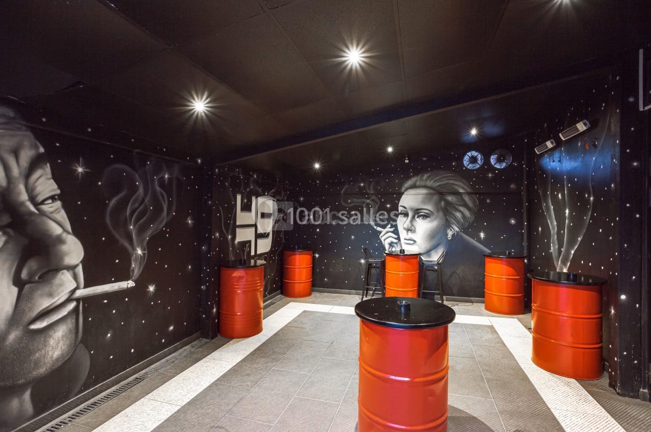 Mur noir décoré de fresques artistiques représentant des visages, avec des tonneaux rouges servant de tables.