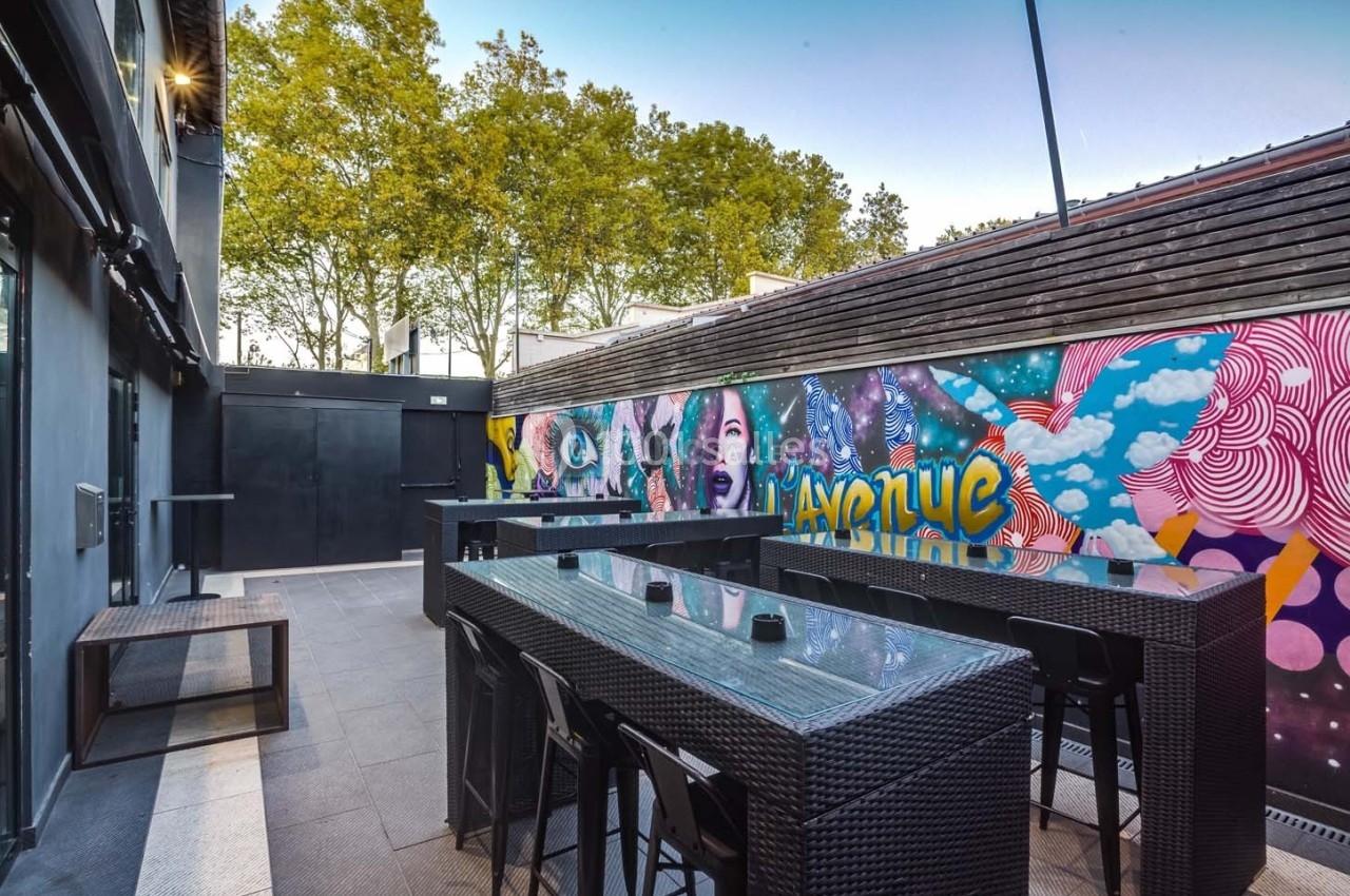 Terrasse extérieure avec tables hautes en rotin noir, murs décorés de fresques colorées et arbres en arrière-plan.