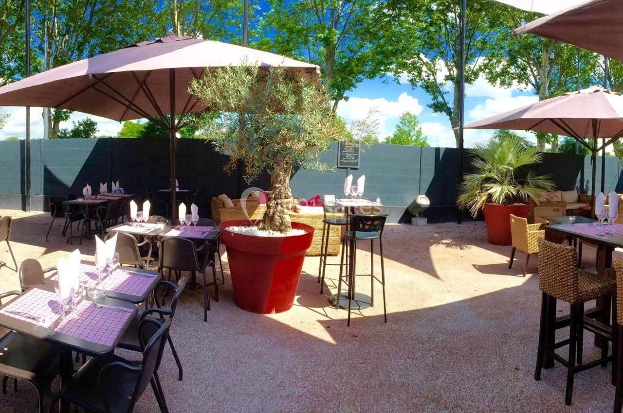 Terrasse extérieure avec tables dressées, parasols, plantes en pot et arbres en arrière-plan.