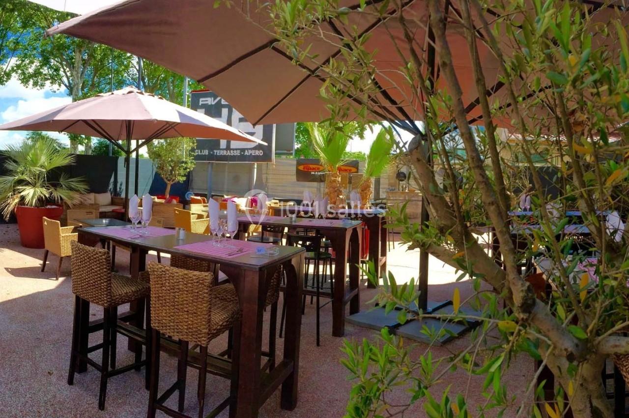 Terrasse extérieure avec tables hautes, chaises en osier et parasols, entourée de plantes et d'arbres.