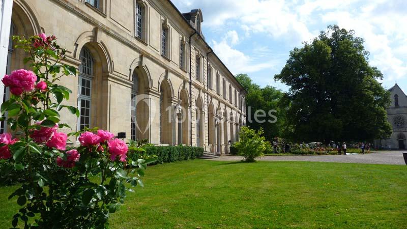 Façade d'un bâtiment ancien avec des arches, entouré d'un jardin fleuri et d'arbres sous un ciel partiellement nuageux.