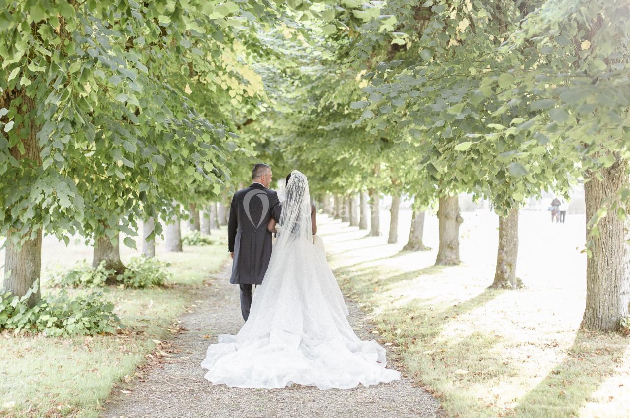 Un couple en tenue de mariage marche sous une allée d'arbres, baignée de lumière naturelle.