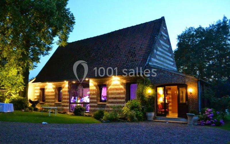Maison en briques avec toit en pente, éclairée par des lumières extérieures, entourée de verdure au crépuscule.