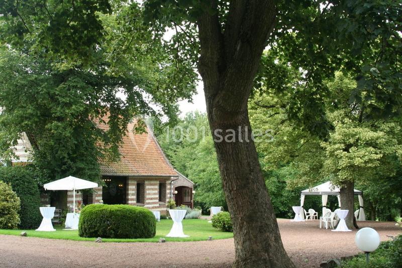 Jardin avec pelouse, arbres, tables hautes et tonnelles blanches près d'une maison en briques et toit en tuiles.