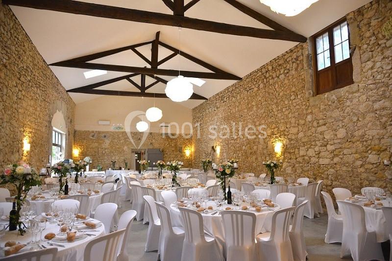 Salle de réception en pierre avec tables rondes dressées, chaises blanches et décorations florales.