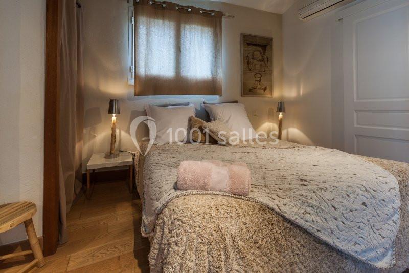 Lit double avec couverture texturée, oreillers blancs, serviettes pliées, lampes de chevet et fenêtre avec rideaux beiges.