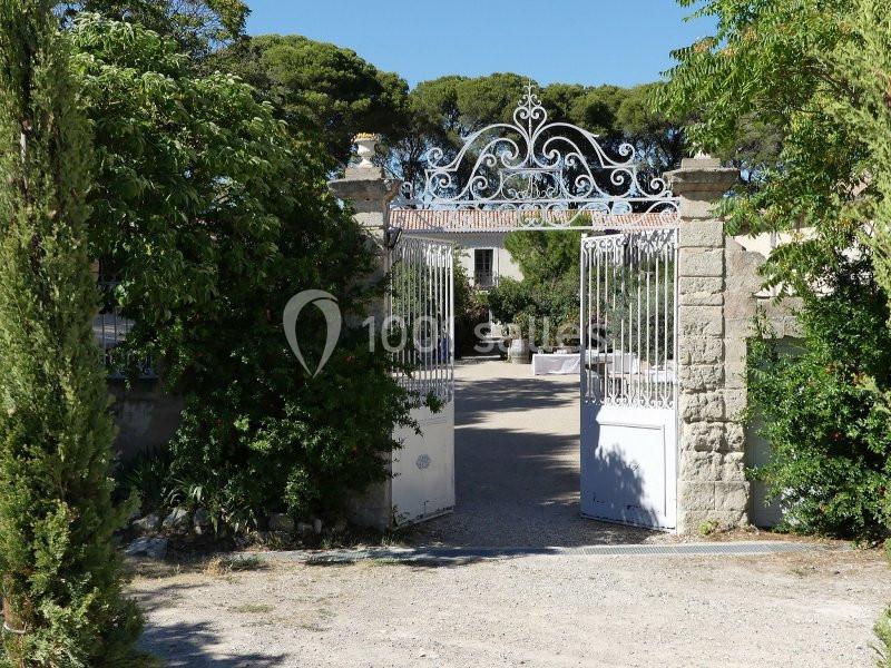 Portail en fer forgé blanc ouvert sur une cour ensoleillée entourée de végétation et de bâtiments en pierre.