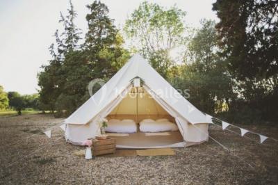 Tente en toile beige installée sur un sol gravillonné, entourée d'arbres, avec des lits aménagés à l'intérieur.