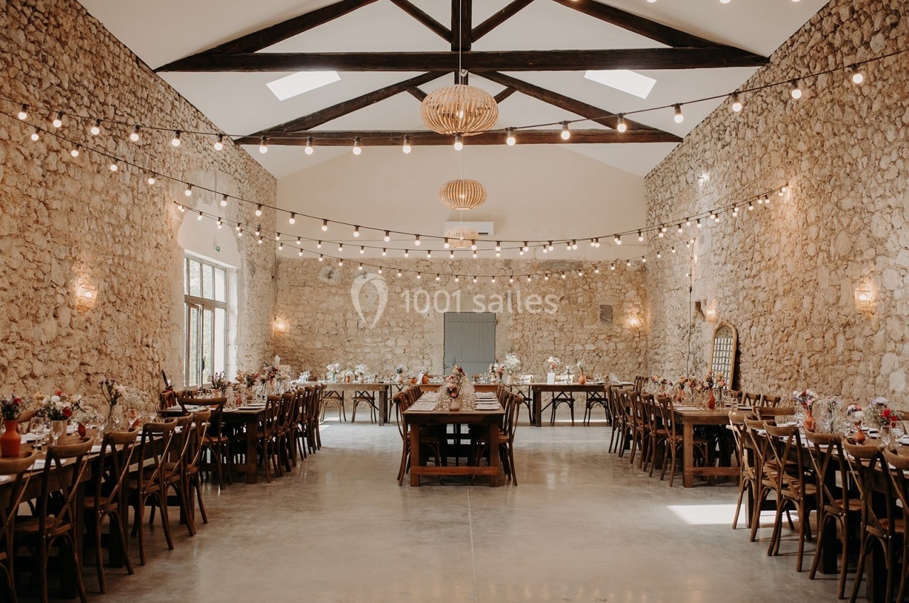 Grande salle en pierre avec tables en bois décorées, guirlandes lumineuses suspendues et lumière naturelle.