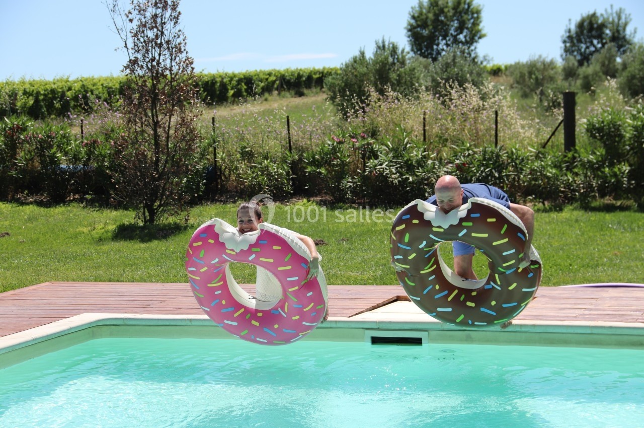 Deux personnes sautent dans une piscine avec des bouées en forme de donuts colorés, dans un jardin ensoleillé.