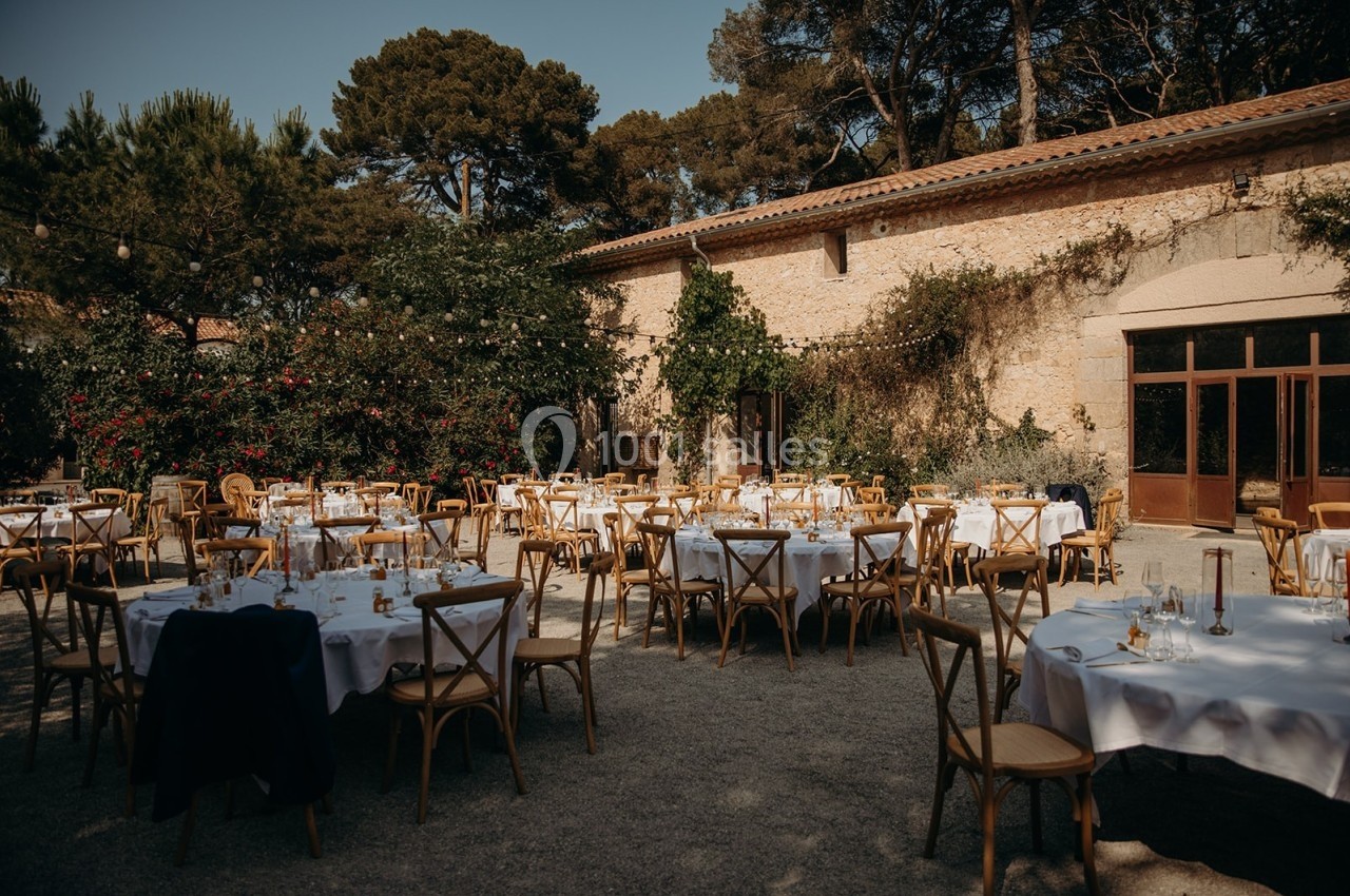 Tables rondes dressées avec nappes blanches et chaises en bois dans une cour extérieure entourée de végétation et de murs en…