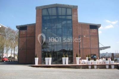 Espace intérieur lumineux avec fauteuils rouges et noirs, tables hautes, plantes et stands d'exposition en arrière-plan.
