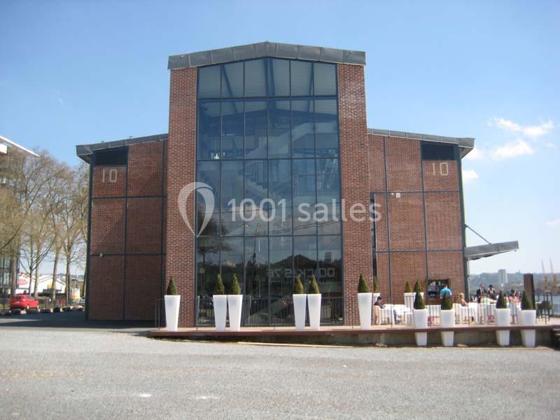 Bâtiment en briques avec une façade vitrée, entouré de pots de fleurs blancs et situé près d'une esplanade.