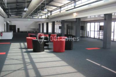 Espace intérieur lumineux avec fauteuils rouges et noirs, tables hautes, plantes et stands d'exposition en arrière-plan.