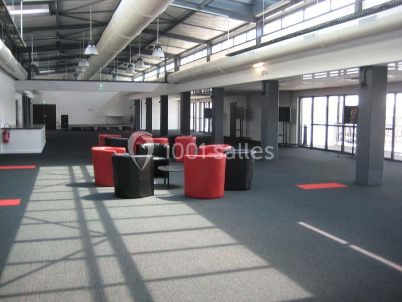 Salle spacieuse avec fauteuils rouges et noirs autour de tables, éclairée par de grandes baies vitrées et un plafond…