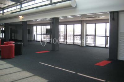 Espace intérieur lumineux avec fauteuils rouges et noirs, tables hautes, plantes et stands d'exposition en arrière-plan.