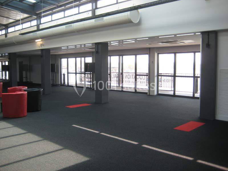 Espace intérieur lumineux avec moquette sombre, baies vitrées et quelques fauteuils rouges disposés près des murs.