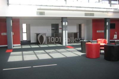 Espace intérieur lumineux avec fauteuils rouges et noirs, tables hautes, plantes et stands d'exposition en arrière-plan.