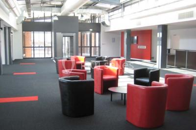 Espace intérieur lumineux avec fauteuils rouges et noirs, tables hautes, plantes et stands d'exposition en arrière-plan.